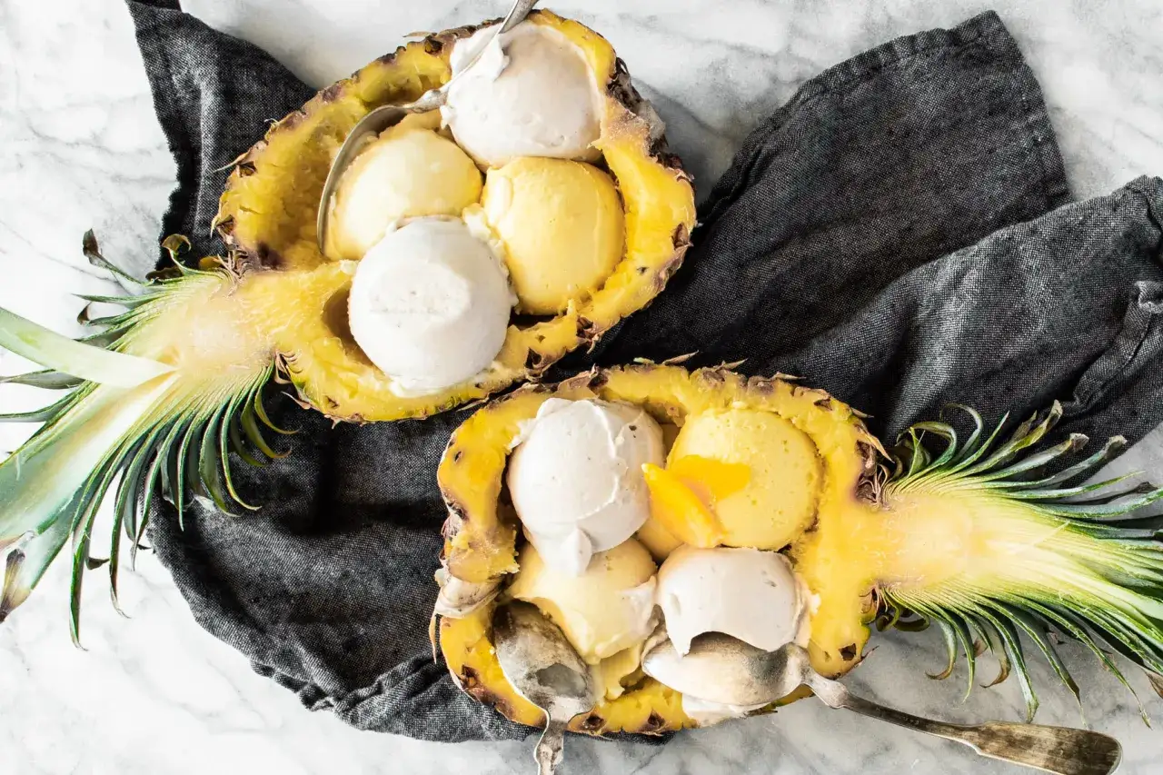 Dos mitades de piña rellenas de helado de coco y otros sabores, servidas con cucharas.