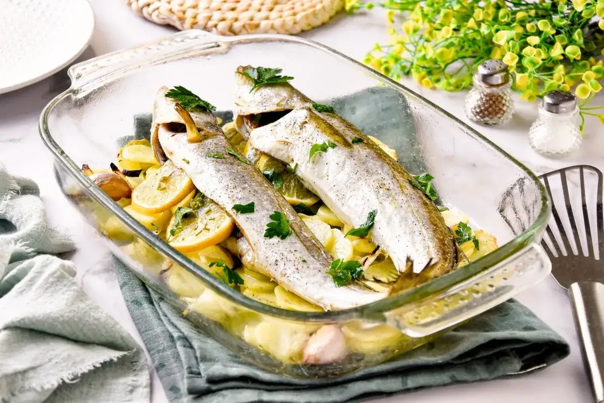 Pescadilla al horno con patatas, limón y ajo, recién salida del horno en una fuente de cristal.