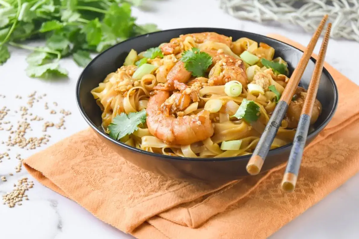 Un taz&oacute;n de fideos de arroz con gambas, adornado con cilantro y cebolleta, listo para ser disfrutado con palillos.