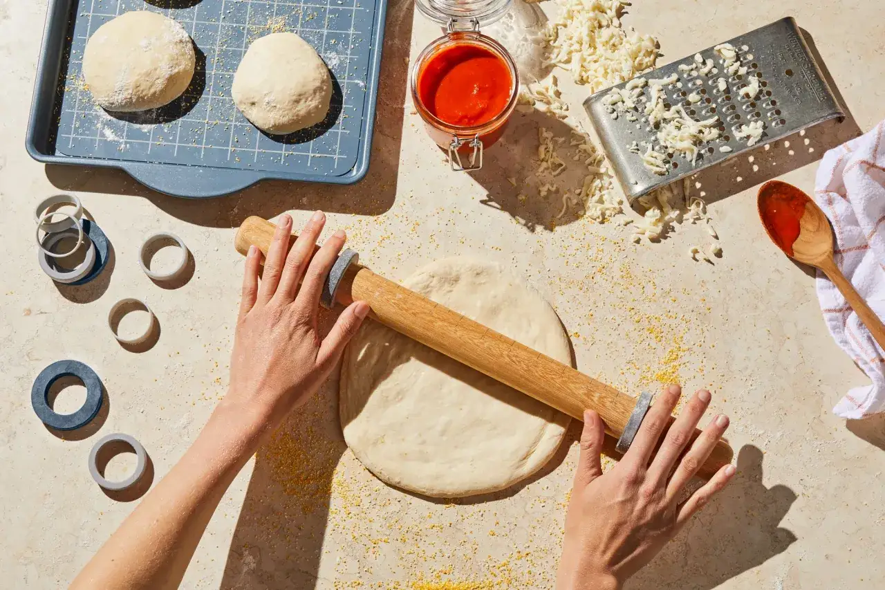 Manos estirando masa de pizza sin levadura con rodillo. Ingredientes como queso rallado, salsa y cortadores circulares listos para usar.