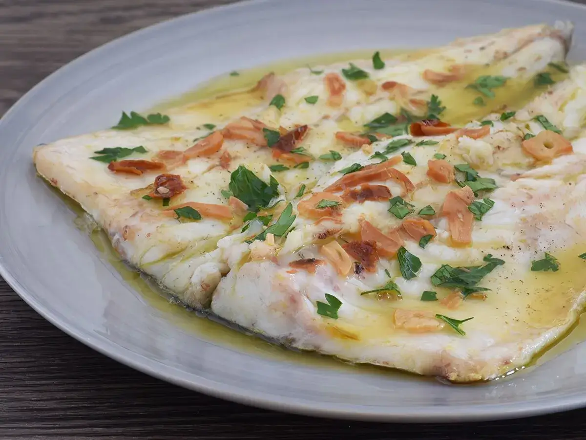 Delicioso lubina a la donostiarra, con láminas de ajo dorado y perejil fresco, servida en una salsa de aceite de oliva.